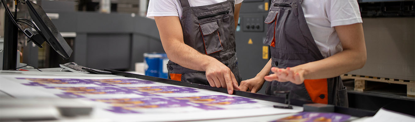eine weibliche Mitarbeiterin und ein männlicher Mitarbeiter in einer Druckerei in Kiel prüfen einen farbigen Großformatdruck auf Fehler
