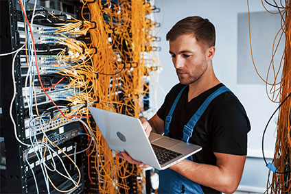 Junger Mann in Arbeitskleidung und mit Laptop arbeitet an der Internetausrüstung und Kabeln im Serverraum.