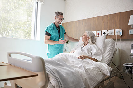 Chirurg besucht ältere Patientin im Krankenhausbett und spricht mit ihr.